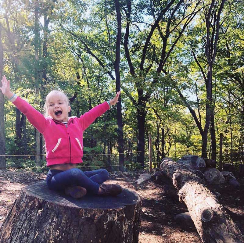 A smiling young girl sitting on a tree stump with her arms up in the air.