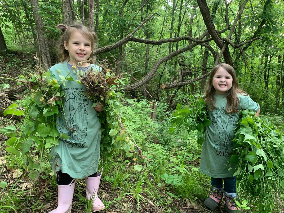 Two young girls each holding a bundle of pulled garlic mustard weeds.