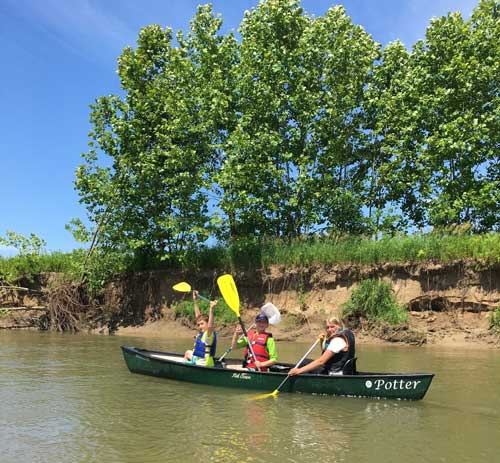 three kids in a canoe holding their paddles up and waving 